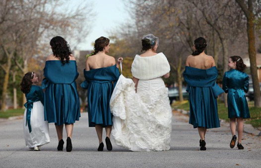 a bride and her wedding party face away from the camera and walk down a road